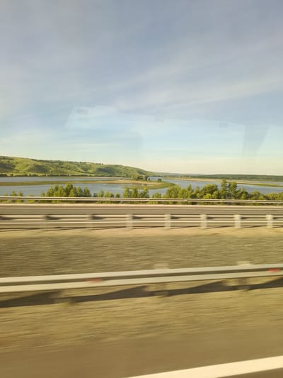 The image captures a scenic view of a wide river with lush green hills in the background. A tree-lined embankment separates the river from a road, which features a white guardrail. The sky is a clear, pale blue with some wispy clouds. The overall impression is one of a tranquil, natural landscape, possibly captured from a moving vehicle due to the slight motion blur in the foreground. The location is identified as Nizhniye Vyazovye, Russia.