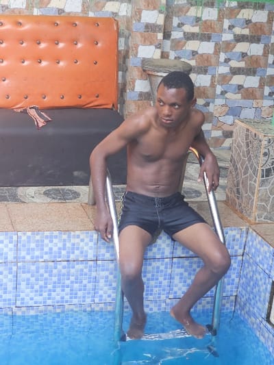 A shirtless man in black shorts is standing in a swimming pool. He is holding onto a metal ladder. The pool is tiled blue. Outside the pool is an orange and black bench and patterned tiles. The location is Asaba, Nigeria.