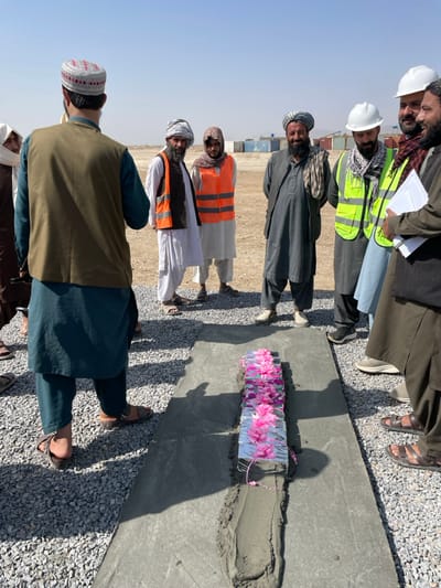 An outdoor, daytime image depicts approximately ten men gathered in a semi-circle around a ceremonial ground-breaking or foundation-laying in Pir Dust, Afghanistan. A rectangular concrete slab, approximately 2-3 feet wide, is laid on a gravel surface. Embedded within a narrow, wet concrete trench on this slab is a silver-wrapped object, adorned with rows of bright pink artificial flowers and a pink beaded string.
The men are dressed in a mix of traditional Afghan attire, including shalwar kameez, turbans, and caps. Some wear modern work attire: two men on the right wear white hard hats and reflective vests (one green, one orange), and one man in the center left wears an orange reflective vest. One man on the far right holds white papers. Many men have beards. The man in the foreground, with his back to the camera, wears a teal shalwar kameez, a brown vest, and a red and white cap, with sandals on his feet.
The environment is an open, arid construction or development site under a clear, bright blue sky. The ground consists of light gray gravel in the immediate foreground, transitioning to light brown, dusty soil in the background. Several shipping containers (blue, white, green) are visible in the distant background. Shadows are cast by the men and objects, indicating direct sunlight.
