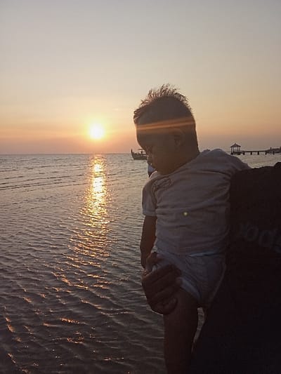 This image captures a sunset scene in Tritunggal, Indonesia. An adult, visible from the right side of the frame with a dark shirt bearing partial white lettering, holds an infant or toddler. The child wears a light-colored short-sleeved top and a light-colored lower garment, with spiky hair and head directed downwards, partially obscured by a lens flare across the upper face. The sun, a bright orange-yellow circle, is low on the horizon, positioned left of center, casting a strong, shimmering reflection across the gently rippled water. In the mid-distance, a dark boat and a pier with a small, roofed structure are silhouetted against the warm-hued sky. The environment is a coastal or lakeside setting.