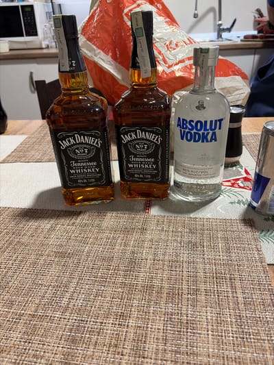 The image shows two bottles of Jack Daniel's Tennessee Whiskey (1 liter, 40% ABV) and one bottle of Absolut Vodka (0.7 liters, 40% ABV) resting on a woven placemat. A can of Red Bull is visible behind the vodka bottle. A red and white plastic bag is partially visible behind the bottles. The setting appears to be a kitchen counter, with a microwave visible in the background. The bottles are likely located in a kitchen in an unknown location. There is no indication of location of Madzherito, Bulgaria.