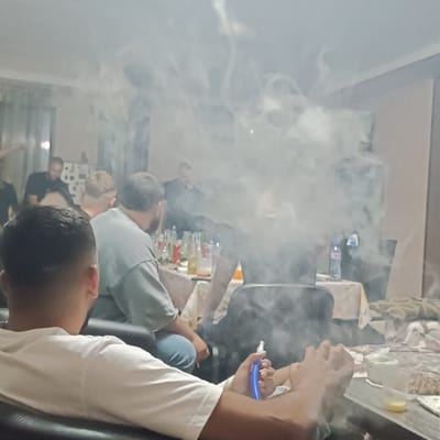 The image depicts an indoor social gathering taking place in Vetren Dol, Bulgaria, likely during evening or nighttime hours due to the artificial lighting. The scene is heavily obscured by dense, white-grey vapor, indicating the active use of multiple hookahs or vape devices.
In the immediate foreground, facing away from the viewer, an individual with short, dark hair and wearing a white short-sleeved t-shirt is seated, their back and right shoulder prominent. This individual holds a blue, plastic hookah or vape mouthpiece in their right hand. Their left arm is visible, and on the dark-colored table in front of them, there is a clear plastic cup containing a yellowish liquid and a white tissue box.
Further into the mid-ground, at least three additional adult males are visible, seated around a table covered with a light-colored, patterned tablecloth. One individual, positioned slightly to the right of the foreground subject, has his back to the viewer, wearing a light grey t-shirt and possessing dark hair and a beard. Another, partially obscured to his left, has lighter-colored, short hair. A third individual is seen through the smoke, wearing a dark shirt. The table is cluttered with numerous bottles of various sizes and colors, including clear, amber, and red bottles, suggesting alcoholic beverages, along with several drinking glasses and cups. A plastic water bottle is also present on a table further back.
In the background, more figures are discernible through the smoke. At least one person in a black t-shirt is standing or leaning towards the left, while another individual is visible further to the right. The walls of the room are light-colored (off-white or light beige), and the ceiling is white. Lighting appears to be from overhead artificial sources, casting a soft, somewhat warm glow. A patterned fabric, possibly a blanket or upholstery, is visible on a surface in the far right background. The overall environment suggests a casual, relaxed social gathering where smoking or vaping is prevalent.