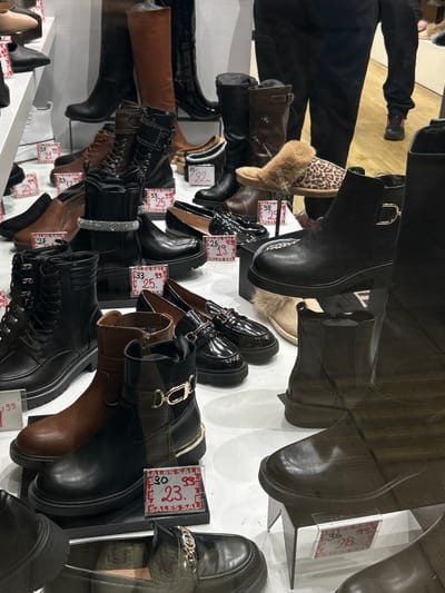 A wide array of women's boots and shoes are displayed for sale in what appears to be a shoe store in Koridallos, Greece. The footwear includes various styles such as ankle boots, knee-high boots, combat boots, loafers, and fur-lined slippers. Dominant colors are black and brown, with some leopard print and tan elements. The shoes are arranged on multiple shelves and stands, creating layers of display. Reflections on a glass surface or mirror add to the visual complexity, showing additional shoes and parts of the store's interior. A person's legs, wearing dark pants and shoes, are partially visible in the upper left corner, suggesting a customer or staff member browsing. The scene captures a shopping environment with numerous price tags indicating ongoing sales. Text on these tags repeatedly features "SALES SALE," alongside various prices like "27," "25," "33," "29.99," "30.99," "23," "38," "29," "36," and "28," likely in Euros. Other numbers such as "199," "292," and "88" are also visible, possibly representing original prices or product identifiers.