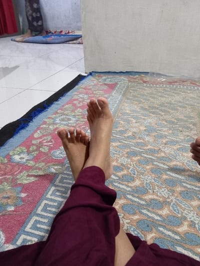 This image depicts an indoor scene in Bulukandang, Indonesia. In the foreground, a person's bare feet are visible, crossed at the ankles, resting on a multi-patterned rug. The individual is wearing dark reddish-purple trousers, covering the legs up to the ankles. The rug displays intricate geometric and floral designs in shades of red, blue, beige, and black. A segment of a plain, light-colored wall is visible on the right side of the frame, extending from the bottom to the top. In the mid-ground, another individual's lower legs and feet are partially visible, wearing dark patterned trousers or a skirt. This second person is standing on a blue prayer mat, which features a white patterned design. The floor beneath and around the prayer mat consists of white square tiles. An additional plain, light-colored wall is visible in the background, behind the second person. A small, translucent plastic object is present on the foreground rug, near the base of the wall on the right.