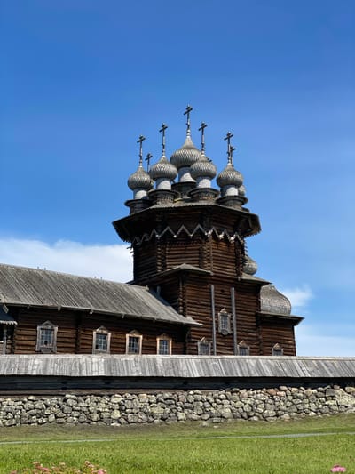 The image shows the Kizhi Pogost, a historic architectural ensemble of churches located on Kizhi Island in the Republic of Karelia, Russia, near the city of Boyarshchina. The main focus is on the Church of the Transfiguration, a wooden church with numerous domes and intricate architectural details. The church is constructed from dark wood and features multiple tiered roofs and ornate carvings. Several silver-colored domes, each topped with a cross, rise from the upper sections of the church. To the left, a lower wooden structure, likely part of the same ensemble, has a gray, weathered wooden roof and several small, multi-paned windows. A stone wall, constructed from rough-hewn stones, runs horizontally across the foreground, separating the buildings from a grassy area with a few pink flowers. The sky above is a vibrant blue with a few scattered white clouds. The overall scene depicts a historic religious site in a natural, rural setting.