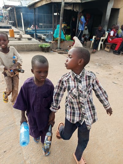 Two young boys are walking on a dirt path in Tumfure, Nigeria. The boy on the left is wearing a light-colored, long-sleeved shirt and tan pants. He is holding a plastic water bottle in his left hand and a dark-colored bottle in his right hand. The boy on the right is wearing a purple tunic and dark pants. He is holding the hands of the boy on the left. In the background, there are market stalls and people. The scene appears to be a busy marketplace.