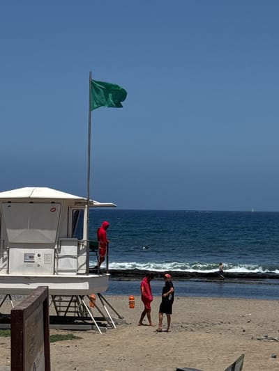 This image depicts a beach scene in Arona, Spain, on a clear, sunny day. In the foreground, a white lifeguard tower stands prominently, with a green flag flying from a pole attached to its side. A person in a red lifeguard uniform is visible on the upper platform of the tower, looking out towards the ocean.
The ocean is a deep blue, with gentle waves breaking on the sandy shore. Further back, two individuals in casual attire are standing on the beach, one in a black t-shirt and shorts, the other in a red top and shorts. Another person is seen lying on the sand near the water's edge.
The beach is sandy and appears to be relatively uncrowded. The time of day is likely midday, given the bright sunlight and clear sky. The overall mood is relaxed and summery. A wooden sign with some text is partially visible in the lower right corner, but the text is not legible. The presence of the lifeguard tower and personnel suggests a public beach where water safety is a priority.
