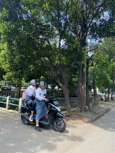 Two young women are sitting on a black scooter. The woman in the front is wearing a white long-sleeved shirt, a white hijab, and a black helmet. The woman in the back is wearing a white long-sleeved shirt, a blue hijab, and a black helmet. They are both looking at the camera. The scooter is parked on the side of a paved road. There is a blue metal railing and a small drainage ditch on the left of the image. On the right side of the image there are trees and bushes. The scene is located in Kecamatan Cimahi Selatan, Indonesia, at the coordinates (latitude: -6.904, longitude: 107.565).