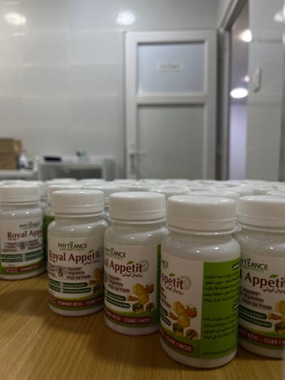 The image displays numerous white plastic bottles, likely containing supplements or medication, arranged in rows on a wooden surface. The bottles are uniformly designed with white screw-on caps and feature green and white labels with text in both French and Arabic. The image is oriented vertically, causing the bottles to appear to be lying on their sides, although they are likely standing upright on a shelf or table.
Each bottle label prominently features "PHYTEANCE LABORATOIRES" at the top, along with the product name "Royal Appetit" and its Arabic translation "روايال آبيتي". Below the product name, key benefits or indications are listed: "Appetit," "Digestion," and "Prise de Poids" (Weight Gain). The main ingredients are also visible, including "Fenugrec & Pollen" (Fenugreek & Pollen) and "Curcuma & Gingembre" (Turmeric & Ginger). A circular logo with "100% Naturel" and a leaf icon is present on the labels. The format and duration of use are indicated as "FORMAT ECO - CURE 1 MOIS" (Eco Format - 1 Month Cure). Additional Arabic text on the labels provides further details, such as "تتكون من أربعة منتجات" (consists of four products), "ينصح باستعماله خلال فترات" (recommended for use during periods), "حالات نقص الطاقة بسبب" (cases of energy deficiency due to), and dosage instructions like "مرتين إلى ثلاث" (two to three times). There are also references to "حبوب الطلع" (pollen grains) and a warning mentioning "و ليس في مكون الأطفال" (and not in children's ingredients).
In the background, a brightly lit room with white tiled walls and what appears to be a white door or window frame is visible. The lighting seems to be artificial, likely from fluorescent fixtures, creating reflections on the smooth surfaces. The overall setting suggests a clean, possibly industrial, storage, or laboratory environment. No specific activity is taking place; the image is a static shot focusing on the stacked products.
