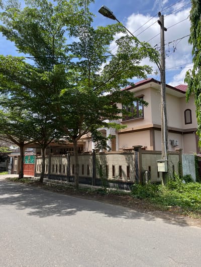 The image shows a street view in Langsa, Indonesia, featuring a modern two-story house with a beige facade and a red tiled roof. The house is partially obscured by several trees with lush green foliage. A decorative fence, constructed with textured stone panels and dark vertical elements, runs along the property line. A brown double gate with decorative cutouts is visible on the left side of the fence. A concrete utility pole with electrical wires and a street lamp stands prominently in the foreground, to the right of the trees. The street in front of the house is paved asphalt, with shadows from the trees cast across it. Dense green vegetation grows along the side of the street and the fence. The sky is partly cloudy. No people or vehicles are visible in the scene.
