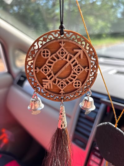 An intricately carved wooden circular charm hangs from a black cord inside a vehicle. The charm features a central design with geometric patterns and symbols, including what appears to be an arrow and a key. Three silver-colored bells and a dark brown tassel with a decorative silver cap are suspended from the bottom of the charm. The background shows the interior of a car, with blurred views of trees and sky through the windshield. The location is not specified as being in Chigiri, Russia; this detail is an assumption.