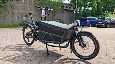 A Riese & Müller electric cargo bike is parked on a hexagonal-patterned paved surface in front of a grey concrete building. The bike is dark gray with a black cargo box covered by a dark green canvas tarp. The bike has a rear rack and a kickstand that is deployed. The front of the bike features a disc brake and a wide tire with visible "SCHWALBE SMART SAM PLUS" text. A tag hangs from the handlebars. In the background, a blue minivan and two dark SUVs are parked. Trees and green foliage are visible behind the vehicles. The scene appears to be outdoors during the day, with bright sunlight casting shadows. The location is indicated as Hamburg, Germany. No people are visible in the image.
