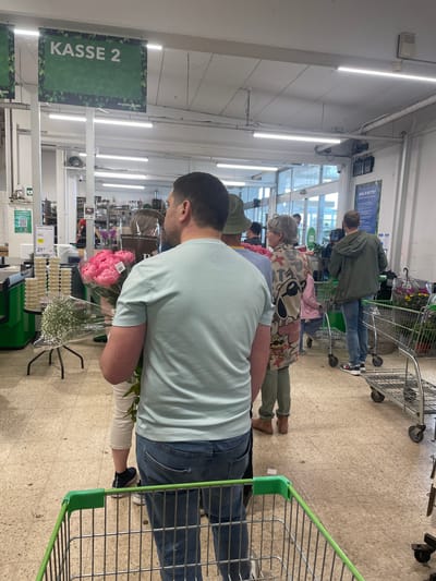 The image captures a queue of shoppers in what appears to be a floral section of a supermarket or department store, likely located in Kolding, Denmark, based on the 'KASSE 2' signage and product prices. The customers are lined up, presumably at a checkout ('Kasse' in Danish). The first person in line, facing away from the camera, is a man holding bouquets of pink peonies and baby's breath flowers, with a green shopping cart in front of him. He's wearing a light blue t-shirt and blue jeans. Several other shoppers are lined up behind him, with their own shopping carts. Above, signage displays the text 'KASSE 2', 'BILLIGBLOMST' and 'SKAL VI BYTTE?', indicating checkout lanes. The environment includes fluorescent lighting, tiled flooring, and displays of various other items.