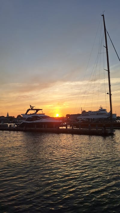 This is a vertical, eye-level, medium shot photograph taken at sunset in Nantucket, Massachusetts, United States. In the foreground, the dark, rippling water of a harbor is visible. Moored to a dock in the midground are several boats. To the left, a modern, dark-hulled yacht with a white superstructure is prominently featured. Further to the right, a larger, white ferry-like vessel is docked. A tall, thin sailboat mast with rigging extends upwards from behind the ferry. The setting sun, a bright yellow orb, is positioned in the center of the frame, casting a warm glow across the sky and reflecting on the water. The sky transitions from a pale blue at the top to a gradient of orange and yellow hues near the horizon, indicative of sunset. In the distance, on the left, a white church steeple can be seen rising above the tree line. The overall atmosphere is serene and tranquil, characteristic of a coastal sunset.