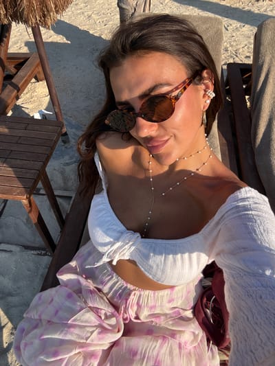 A woman is lying on a wooden lounge chair on a beach. She is wearing a white off-the-shoulder top, a pink and white patterned skirt, and sunglasses. She has a pearl necklace and pearl earrings. The sun is shining, casting shadows on the sand and the lounge chair. The scene suggests it is a sunny day in Bodrum, Turkey, likely during the daytime, and the woman appears relaxed. There is a straw-like object in the lower left corner, possibly part of a beach umbrella or mat. The overall ambiance is that of a leisure day at the beach.