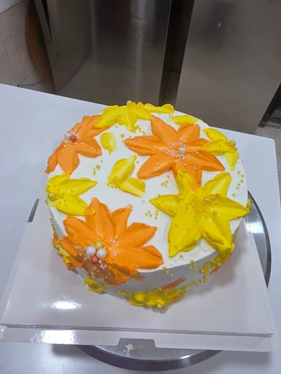 A round cake with white frosting is decorated with orange and yellow buttercream flowers and leaves. Several small yellow and white pearl-like candies are scattered on top and around the flowers. The cake is sitting on a white cake board, which is on a silver turntable. The background shows a metallic surface, possibly part of kitchen equipment. The scene is brightly lit. The location is identified as Dibba Province, Oman.