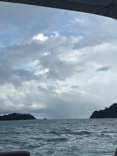 The image depicts a seascape with a stormy sky and rough water. The perspective is from a boat, indicated by the dark frame on the left. In the sky, there are dramatic clouds and a faint rainbow. A small cluster of pink objects, possibly kites or balloons, are visible near the rainbow. On the right side of the frame, the choppy water is detailed, and a dark, tree-covered landmass with a building and a structure resembling a lighthouse is visible in the distance. The overall mood is somber, suggesting inclement weather, possibly late afternoon or evening given the lighting. The location is identified as Ratsada, Thailand.