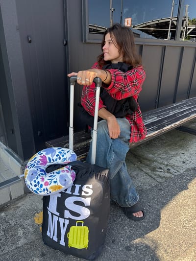 A young woman with long, dark hair is sitting on the ground next to a black suitcase. She is wearing a red and black plaid shirt, blue jeans, and black sandals. The suitcase has a floral-patterned neck pillow and a black bag with "THIS IS MY..." printed in large white letters, along with a yellow suitcase icon. The woman is holding the handle of the suitcase, which has extended silver poles. She is looking down and smiling, with several rings on her fingers. The scene appears to be outdoors, with concrete ground and a dark building in the background. The lighting suggests it is daytime, possibly late afternoon due to the long shadows cast. The overall mood seems relaxed and perhaps anticipatory of travel.