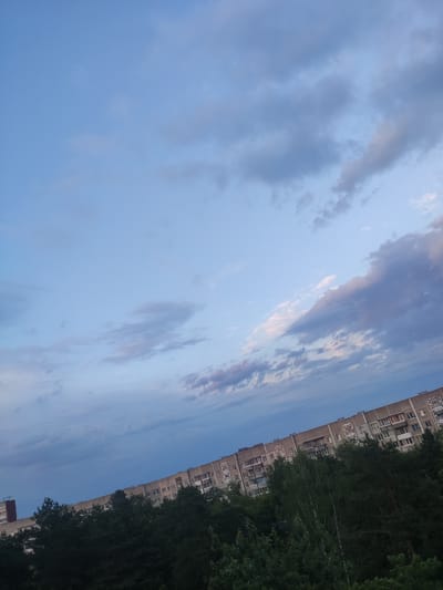 A wide-angle, full-frame photograph captures a suburban residential building and trees under a cloudy sky in Navapolatsk, Belarus. The building, a multi-story apartment complex, stretches diagonally across the frame from the lower right to the upper left. It is constructed of light-colored material and features numerous windows, balconies, and architectural details. In the foreground, a dense canopy of dark green trees extends across the lower portion of the image, obscuring the base of the building. The sky above is a mix of light blue and gray clouds, with hints of pinkish hues, suggesting either dawn or dusk. The overall lighting is soft and diffused, characteristic of overcast conditions. No people are visible in the frame. The scene depicts a common urban or suburban landscape.