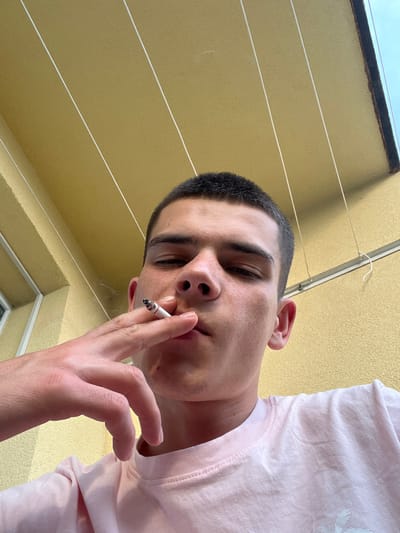 The image features a close-up, low-angle shot of a young person holding a lit cigarette to their mouth. The person has short, dark hair and is wearing a light pink t-shirt. The background is a textured, light yellow wall with horizontal white lines running across it, possibly clotheslines. A dark metal bar with a white hook is also visible near the top of the frame, suggesting an outdoor setting, possibly a balcony or patio. The lighting suggests it is daytime, and the overall mood is casual. There is no visible text in the image. The location context provided is Sopot, Bulgaria.