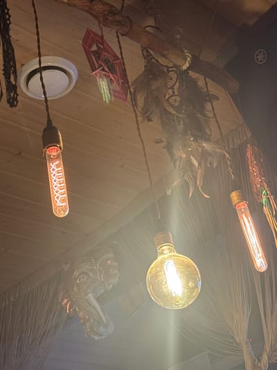 The image depicts an indoor scene with a rustic aesthetic, likely a bar or restaurant interior, in Krasnaya Polyana, Russia. The ceiling is constructed of wooden planks. Several decorative elements are visible, including a circular white light fixture, a red woven geometric ornament, and a collection of feathers arranged as a dreamcatcher. Hanging lights are a prominent feature: one is a tubular bulb with a visible filament, another is a round bulb emitting a warm glow, and a third is another tubular bulb with a similar filament. A carved wooden artifact, possibly an animal head, is mounted on the wall or ceiling. The lighting is warm and somewhat diffused, creating a cozy atmosphere. No people are present in the image. The overall impression is one of a thoughtfully decorated, dimly lit space with a focus on natural and handcrafted elements.