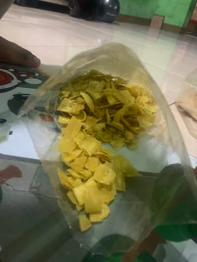 A clear plastic bag containing a large amount of fried banana chips sits on a tiled floor. The bag is partially open and the chips are visible. The floor appears to be made of light-colored tiles. There is a partial view of a person's foot in the lower left corner of the image. The image was taken in a home in Lagadar, Indonesia. The GPS coordinates provided are -6.928 latitude and 107.535 longitude.