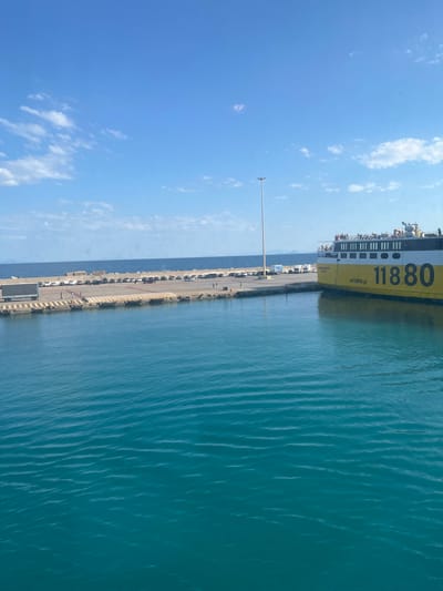 A yellow and white ferry, identified as "11880" and bearing the website "vrrisko.gr", is docked at the port of Kyllini, Greece. The ferry has people standing on its upper deck. Adjacent to the ferry is a pier where numerous cars are parked. A large semi-trailer truck is also visible on the pier, along with several pedestrians. The water in the foreground is a vibrant turquoise color with gentle ripples. In the background, a calm sea meets a shoreline with mountains or hills in the distance under a clear blue sky with scattered white clouds. A tall light pole stands on the pier. The overall scene depicts a busy port environment on a clear day.