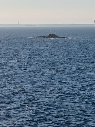 The image shows a body of water, likely the sea, with a small rocky island in the distance. On top of the island, there appears to be a lighthouse or a similar structure with a red top and a white pole. The water is choppy, indicating moderate waves. In the far distance, a hazy landmass with several tall structures, possibly industrial smokestacks or communication towers, is visible. The sky is clear and pale. The overall scene suggests a maritime environment, possibly near a coastline. Without further information, the specific location in Helsinki, Finland, cannot be determined. There are no people, animals, or other discernible objects in the immediate vicinity of the island or in the water.