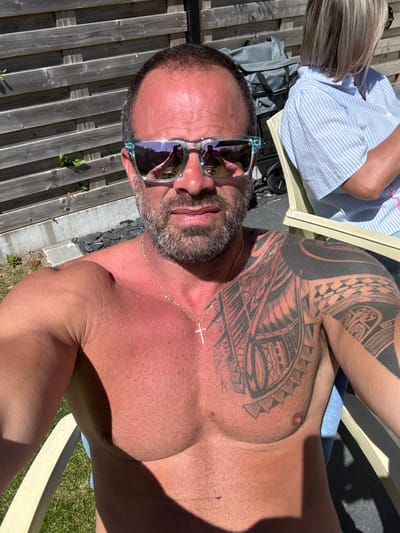 A medium-angle, high-resolution daytime outdoor selfie shot shows a shirtless man in sunglasses with a beard and patterned shoulder/chest tattoos, wearing a gold cross necklace, seated in a light-colored chair next to a woman in a striped shirt and a grey backpack, alongside a wooden fence and green grass. The photo was taken in Bredene, Belgium.