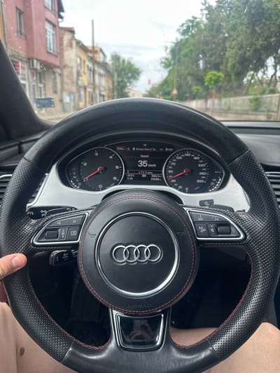 The image shows a close-up view of an Audi steering wheel and dashboard from the driver's perspective. The steering wheel is black leather with red stitching and features the Audi logo in the center. Several buttons are visible on the steering wheel, including controls for navigation, audio, and cruise control. The dashboard displays a speedometer with a needle indicating a speed of 35 km/h, as well as a digital display showing other vehicle information. The background shows a blurred view of buildings and trees through the car window, suggesting the vehicle is in motion in an urban or suburban environment. The light conditions suggest it is daytime. The scene is from inside a car, likely in Kazanlak, Bulgaria, given the location context. There are no discernible people clearly visible, though a hand is partially visible holding the steering wheel. The overall mood is neutral, focusing on the car's interior and its operation. Visible text includes "SPEED", "km/h", "NAV", "AIRBAG", and various symbols and numbers on the dashboard.