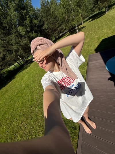 A young woman is depicted in a full-body selfie, standing barefoot on a dark brown slatted wooden deck under bright, direct sunlight. She is wearing an oversized white t-shirt with red and blue text, partially visible, that includes "APRESS INSTRUCTIO," "HOTLINE," "3-777-3571," and "Ski Bum Wisdom." A pink terry cloth towel is wrapped around her head. Her left hand is raised to shield her eyes from the sun, casting shadows on her face. Her right arm is extended, holding the camera. The background consists of a lush green grassy lawn that gently slopes upward, populated by numerous tall trees, including distinct birches and dense conifers, under a clear blue sky. Strong shadows are cast across the deck and grass, indicating intense sunlight. A small, partially visible blue object is situated on the far right of the deck. This scene is located in Копнино, Russia.