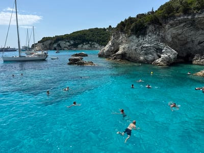 In the city of Ozias, Greece, a group of people are swimming and enjoying the clear turquoise waters of a cove. The scene is set against a backdrop of rugged, white-streaked cliffs covered in lush green vegetation. Several sailboats are anchored in the water, with one prominent white sailboat in the foreground, its mast reaching towards the clear blue sky. The water is calm and inviting, with visible ripples on its surface. A few people are actively swimming, while others appear to be floating or relaxing. Some rocks are visible breaking the surface of the water. The overall impression is one of a serene and picturesque coastal destination.