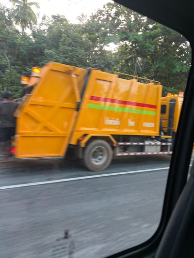 A blurred image taken from inside a vehicle shows a bright orange garbage truck driving on a paved road. The truck has green and red stripes on its side, and orange flashing lights on top. There are trees and foliage in the background. The image appears to be taken in Ban Khao Hua Chang, Thailand, as indicated by the text on the truck, which is in Thai script.