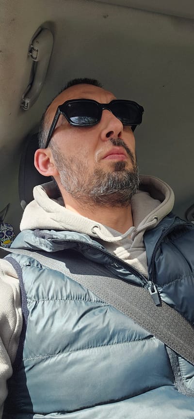An upright-oriented image features a man in a car, likely a passenger. He is seen from the chest up, wearing a light grey or beige hooded sweatshirt layered under a teal or blue-green puffer vest. A grey seatbelt diagonally crosses his chest from his left shoulder.
The man has a beard and mustache, showing noticeable streaks of grey hair. He wears black, rectangular sunglasses that obscure his eyes. His expression appears neutral and relaxed. Bright sunlight illuminates the scene, suggesting it is daytime and possibly sunny outside.
The car's interior is visible, with a light grey or beige fabric headliner on the ceiling. An overhead grab handle, matching the interior color, is mounted above and to the right of his head. No visible text is present on his clothing, accessories, or the car's interior. The setting is a casual car journey, taking place in Norwich, United Kingdom.