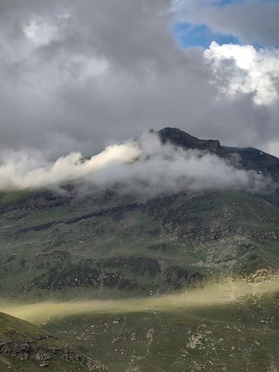 This is a vertical, medium-shot photograph of a mountain landscape in Gramphu, India. The foreground shows a steep, rocky incline covered in sparse green grass. The midground features a large, undulating mountain slope, also covered in green vegetation and scattered rocks. A band of bright sunlight illuminates a portion of this slope, creating a vivid green and yellow stripe across the otherwise muted greens and grays. The upper portion of the mountain is rugged and dark, with patches of snow visible. Clouds obscure the peak, with some white, fluffy clouds near the summit and darker, stormier clouds filling the upper sky. A sliver of blue sky is visible between the clouds. No people or man-made objects are present in the image.