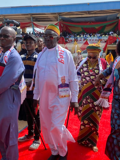 The image shows a group of people in Takau, Nigeria, likely at an event. The foreground features several individuals dressed in traditional attire, wearing sashes that seem to commemorate the appointment of "GENERAL CG MUSA (rtd) OFR". One man wears a white robe and a matching sash with the words "SERVICE" and "ating" visible. Another wears a patterned robe and hat. A red carpet covers the ground. In the background, spectators are visible, seated in rows under a structure decorated with flags or banners in red, green, and yellow. The sky is light blue.