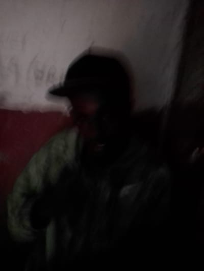 The image, captured in Wirana, Indonesia, presents a blurry, low-light scene featuring a single individual from the chest up. The person wears a dark baseball cap and a light-toned, possibly greenish-brown or olive-colored, long-sleeved garment. The individual's facial features are largely indistinct due to deep shadow and motion blur. The immediate background comprises a two-toned wall: an upper section of light grey or off-white hue, exhibiting faint, dark, illegible markings resembling graffiti or symbols; and a lower section painted a distinct dark red or maroon. No specific actions or interactions are discernible due to the image quality and lighting conditions.
