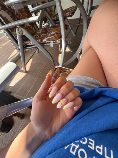 A close-up shot focuses on a person's hand with long, almond-shaped nails, featuring a light pink and pale yellow French tip design. One nail is adorned with a small, shimmering flower embellishment. The hand rests on a blue garment, possibly a shirt or jacket, with white Cyrillic lettering visible on it. The text appears to say "ПРЕСС ОД". The background shows a wooden deck, metal chair frames with floral-patterned fabric, and sunlight casting shadows. The scene suggests a casual outdoor setting, likely during the day given the strong sunlight and shadows. The overall impression is one of relaxation or leisure. The location is specified as Bulgarovo, Bulgaria.