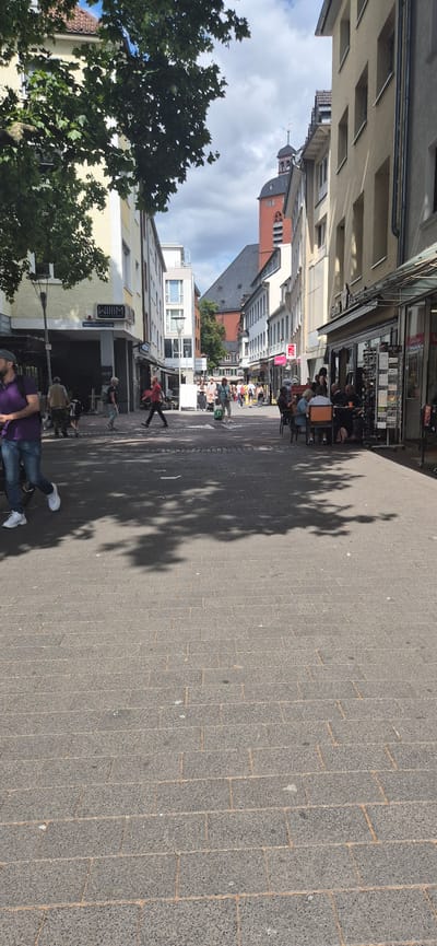 The image captures a view down a pedestrian street in Mainz, Germany. The street is paved with cobblestones and lined with multi-story buildings housing shops and cafes. On the left, a person in a purple shirt and jeans walks away from the camera, with their left foot in focus. Further down the street, several other individuals are visible walking, some with shopping bags, and others pushing strollers. A group of people are seated at tables outside a cafe on the right side of the street. In the background, a church tower with a distinctive red brick spire is visible, and the sky is partly cloudy. The scene appears to be a typical sunny day in a European city, with a mix of locals and tourists enjoying the atmosphere.