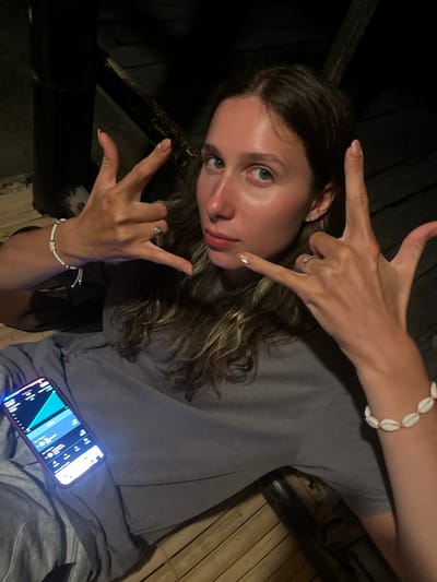 A young woman with long, wavy brown hair is seated and looking at the camera, holding up her hands in a "rock on" gesture. She is wearing a gray t-shirt and a white bracelet with small shells. There is a ring on her right ring finger and another on her left ring finger. A smartphone is visible in her lap, displaying a screen with charts and text. The background appears to be made of wooden planks, possibly a deck or floor, with dark vertical supports. The lighting is dim, suggesting an outdoor evening or dimly lit indoor setting. The location is stated to be in Kertasari, Indonesia, though the image itself does not provide specific visual cues to confirm this.