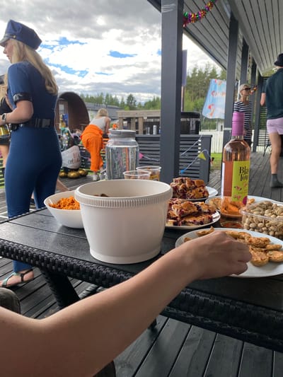 This image depicts a gathering outdoors on a cloudy day, likely in Joensuu, Finland. In the foreground, a person in a blue uniform with a hat and belt is visible from the side, with their arm extended towards a table laden with food and drinks. The table is set with white plates containing pastries, nuts, and what appears to be some sort of savory snack. A bottle of rosé wine with a pink label, reading "Black Tower Pink Grape," is also present. A large white mixing bowl and a smaller bowl filled with orange-colored food are on the table. In the background, other people are present. One person is wearing orange overalls, and another is seen from the torso up. Decorations, including colorful streamers and balloons, suggest a festive occasion. The scene is set on a wooden deck, likely a patio or veranda, with a black wicker chair visible. The overall atmosphere suggests a casual outdoor event or party.