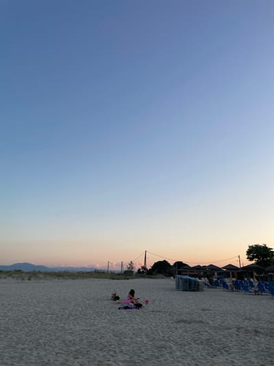 The image depicts a sandy beach in Greece during what appears to be late afternoon or early evening. The sky is a clear gradient of blue to pale orange, indicating the setting sun. In the foreground, the sand is textured with footprints and small ripples. To the right side of the frame, a row of blue lounge chairs and straw-covered umbrellas are neatly stacked or set up, suggesting a resort or public beach area. In the middle ground, a person in a pink dress is sitting on a blanket on the sand, appearing to be relaxed. There are also bags and a small stuffed animal nearby. Further in the background, there are trees, some vegetation, and utility poles with wires. In the far distance, mountains are visible against the horizon, with soft, pinkish clouds. The overall scene conveys a peaceful and serene atmosphere.