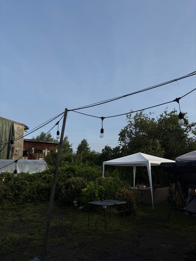 An outdoor scene at dusk in Jāņupe, Latvia, shows a backyard area with a white pop-up canopy tent set up over a table with chairs. String lights are suspended from wires overhead, with several bare bulbs visible. To the left of the canopy is a greenhouse or similar structure. A wooden pole supporting some of the wires stands in the foreground. The ground appears to be muddy grass. Trees and dense green bushes are abundant in the background, along with a portion of a building. A swing set is partially visible on the right side of the frame. The sky is a clear, pale blue, indicating either early morning or late evening.