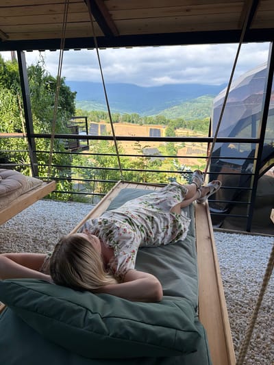 A blonde woman with her arms behind her head is lying on a suspended wooden lounger covered with a green cushion. She is wearing a floral print dress and white sandals. The lounger is positioned on a balcony or outdoor area covered with white gravel. In the background, a picturesque view of lush green mountains under a cloudy sky is visible. The scene appears to be set in a natural environment, possibly a resort or private residence in Nizhnyaya Shilovka, Russia, given the architectural style and the landscape.