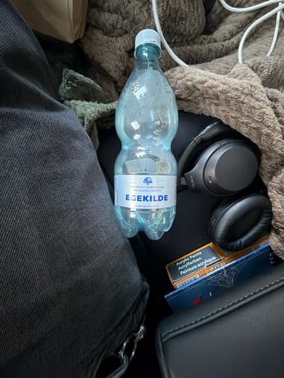 The image shows a close-up, overhead view of personal items resting on what appears to be car seats or a similar upholstered surface. In the center of the frame is a clear plastic bottle of "EGEKILDE" mineral water, with a blue and white label. To the right of the water bottle are a pair of black Sony headphones. Below the headphones and partially obscured by them is a small box labeled "Acrylic Paints," with multilingual text including "Acrylpaints" and "Peinture acrylique." A brown, fuzzy blanket or throw is draped over the top and right side of the scene, and a section of dark, textured fabric, possibly denim jeans, is visible on the left. The lighting suggests an indoor or vehicle setting. The scene is static, with no observable actions or interactions. The location in the city of Ödsmål, Sweden, is not discernible from the visual elements present in the image.