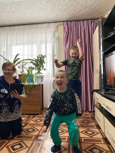 A photograph depicts a scene in Bely Klyuch, Russia. An elderly woman wearing glasses and a patterned blue and white shirt is sitting on the floor and holding a tablet or large smartphone. Two young boys are positioned behind her. One boy is wearing a dark green t-shirt. The other boy wears a patterned shirt and green pants. A large potted plant stands to the left of the woman, positioned in front of a window covered with white sheer curtains. A brown cabinet sits behind the woman. The floor is tiled with brown and beige patterns. Above the boys, a purple curtain is visible below what appears to be a television and storage units mounted on the wall. The interior space has patterned white wallpaper.