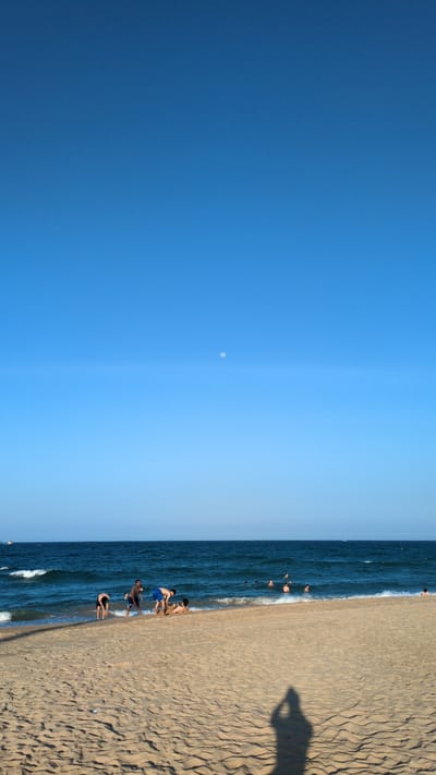 The image depicts a beach scene with the sky taking up approximately half the frame. The sky transitions from a darker blue to a lighter shade. A small, pale circle, possibly the moon, is visible in the lighter blue section. The lower half of the image shows a sandy beach meeting the ocean. People are seen in the water and on the shoreline. The sand displays textured patterns and a shadow of a person, presumably taking the picture, is cast onto it. The water is dark blue with small waves. The location appears to be a beach in Thành phố Tuy Hòa, Vietnam. People are interacting near the water's edge.