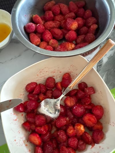 The image displays a close-up overhead view of two bowls filled with strawberries. The bowl on the left is a stainless steel bowl containing whole strawberries. To its left is a small white bowl with a yellow liquid, likely juice or syrup. The bowl in the foreground is a white, shallow bowl filled with chopped strawberries. A silver knife is positioned on the left edge of this bowl, and a spoon with a wooden handle is resting inside, partially submerged in the strawberries. The background shows a speckled countertop and a dark surface. There are no people visible in the image. The scene depicts food preparation, likely for a dessert or jam. The location is not specified in the image or crops provided.