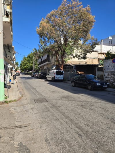 This is a ground-level, eye-level, slightly low-angle, wide shot of a street in Athens, Greece, on a clear, sunny day with a bright blue sky. The scene depicts a narrow street with buildings on either side and parked cars.
On the left side of the street, there's a building with a green-painted section at the front and a balcony with a railing. Further down the sidewalk, there are some trees and parked cars. The sidewalk appears to be paved with light-colored tiles. A black trash can is visible next to a blue recycling bin.
In the center of the street, there is a large tree with a thick trunk and a full canopy of leaves, some of which appear to be turning yellow and brown. The street itself is paved with asphalt and shows signs of wear and tear, with oil stains and cracks visible. There's a metal manhole cover in the road.
On the right side of the street, there are parked cars, including a dark blue Volkswagen Golf and a white van. The buildings on this side are varied in style, with some appearing older and somewhat weathered. One building has a large sign with Greek text. Further down the street, there are more parked cars and trees. The scene conveys a sense of a typical urban street in Greece.