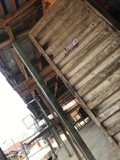 The image depicts an outdoor market scene in Akure, Nigeria, characterized by a network of weathered, makeshift structures viewed from a low, tilted perspective. In the upper right foreground, a corrugated panel, possibly tin or wood, shows significant wear, dirt, and light-colored streaks; affixed to it is a rectangular sticker featuring the words "Bad Spare Parts" and illustrations of vehicle components. Dominant structural elements include multiple rough-hewn wooden beams and at least one prominently featured vertical beam painted a faded light green/teal, all appearing aged and structurally supporting various overhead corrugated metal sheets and wooden planks that form roofs and ceilings.
Through the gaps and beneath these structures, a street or open market area is visible in the mid-ground and background. One individual, wearing a red shirt, is seen walking away from the viewer on what appears to be a dirt or uneven paved road. Further down the street, a yellow tricycle (keke Napep) is partially visible, indicating local transportation. Other distant figures and additional rudimentary market stalls or buildings line the street. The ground immediately beneath the visible structures appears to be concrete or packed earth. The scene is illuminated by natural daylight, with areas under shadow from the overhead coverings.